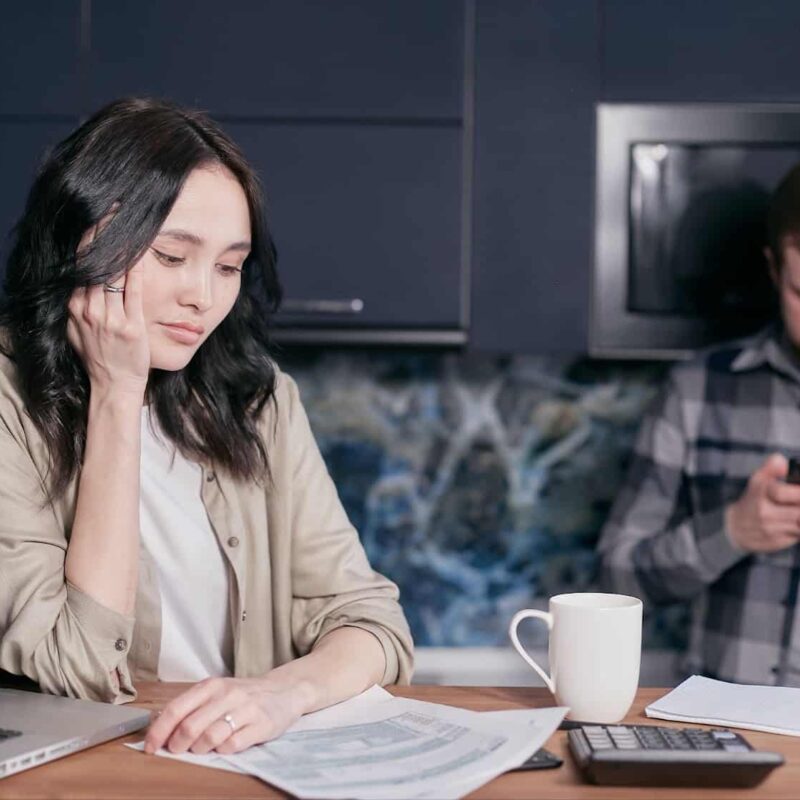 Woman at table looking sadly at a group of papers, while a man in the background texts | New Direction Family Law