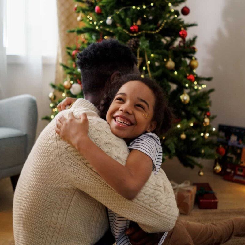 A young girl hugging her father next to a Christmas tree. | New Direction Family Law