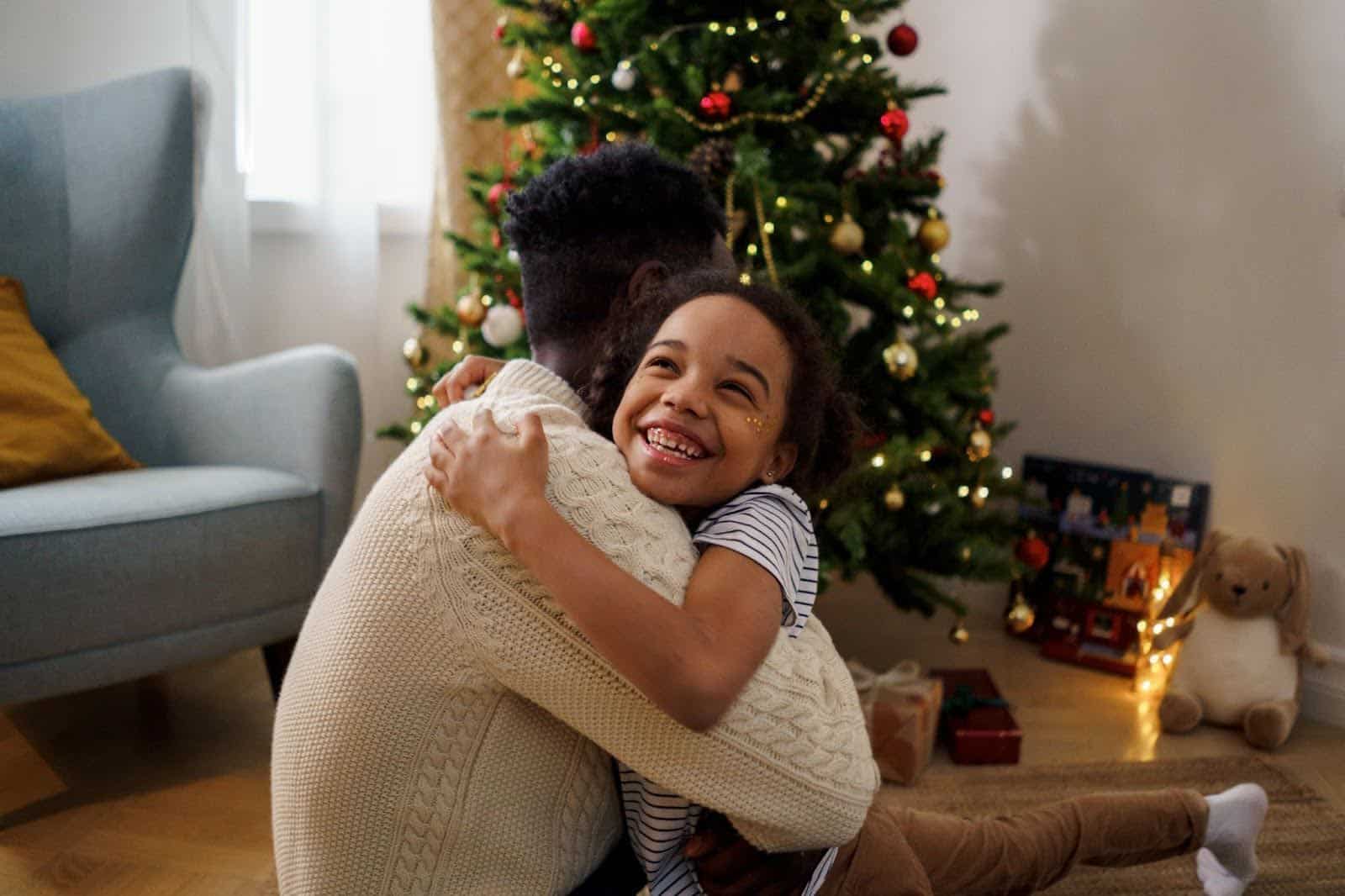 A young girl hugging her father next to a Christmas tree. | New Direction Family Law