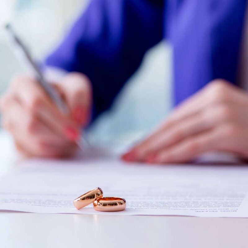 Two rings sitting on a prenuptial agreement as a person signs the document.