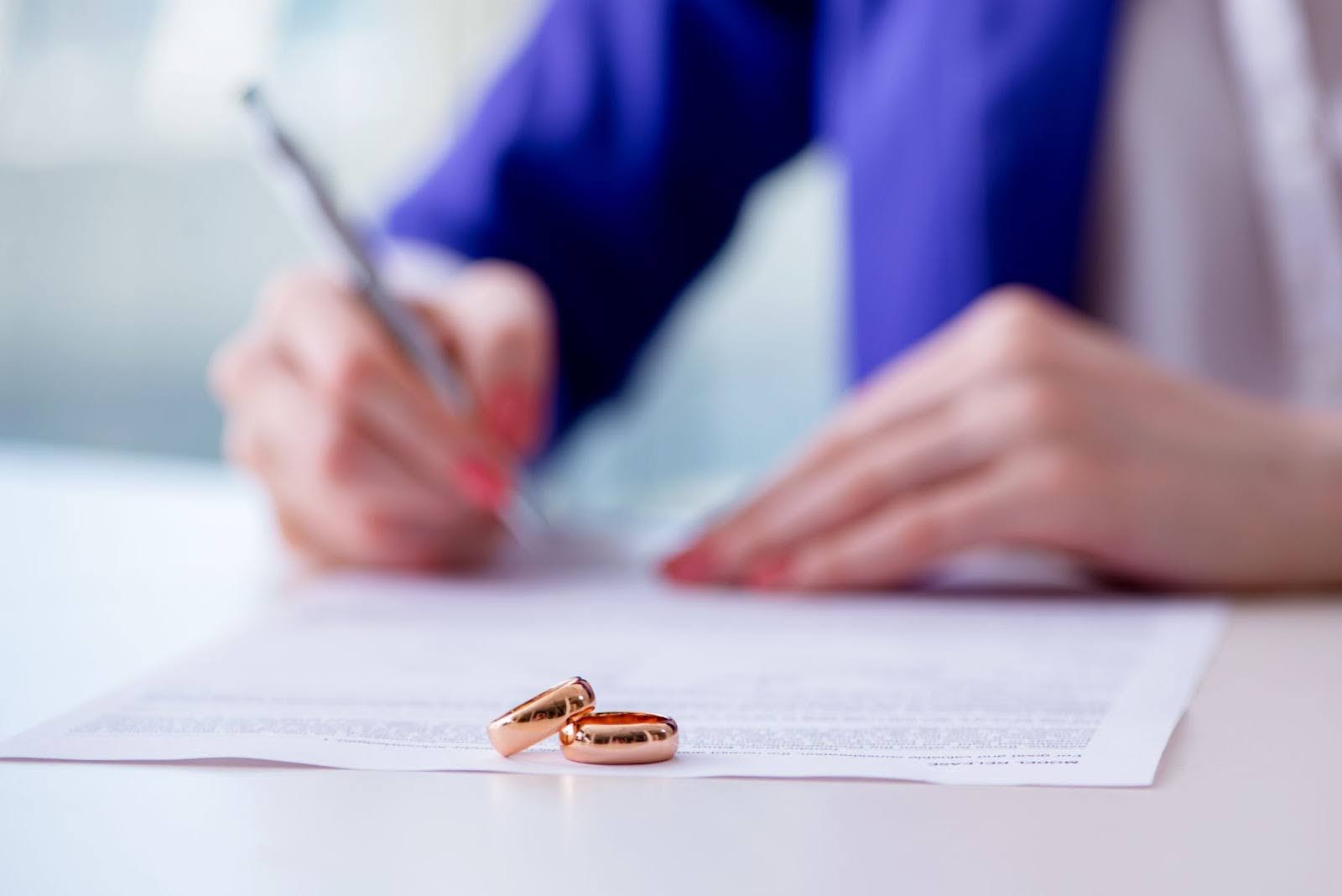 Two rings sitting on a prenuptial agreement as a person signs the document.